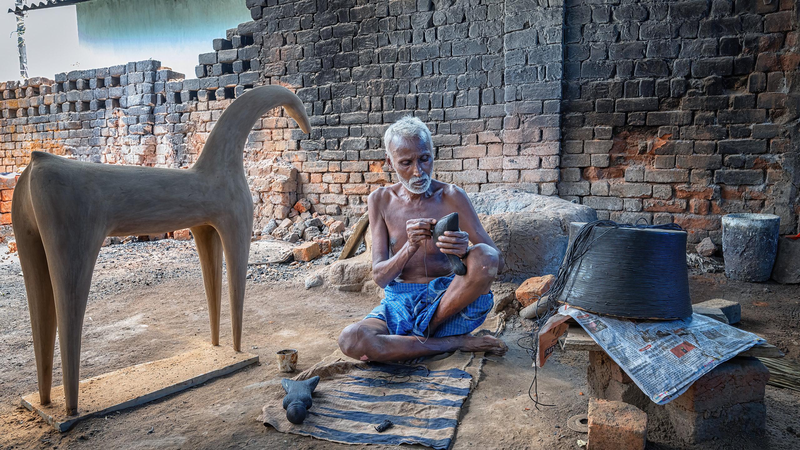 An artisan winding wax thread onto a small form, seated between a standing clay core and a coil of drawn wax.